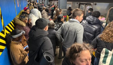 Photo shows a large crowd of people waiting to board a train on a platform partially obstructed by a blue construction wall.